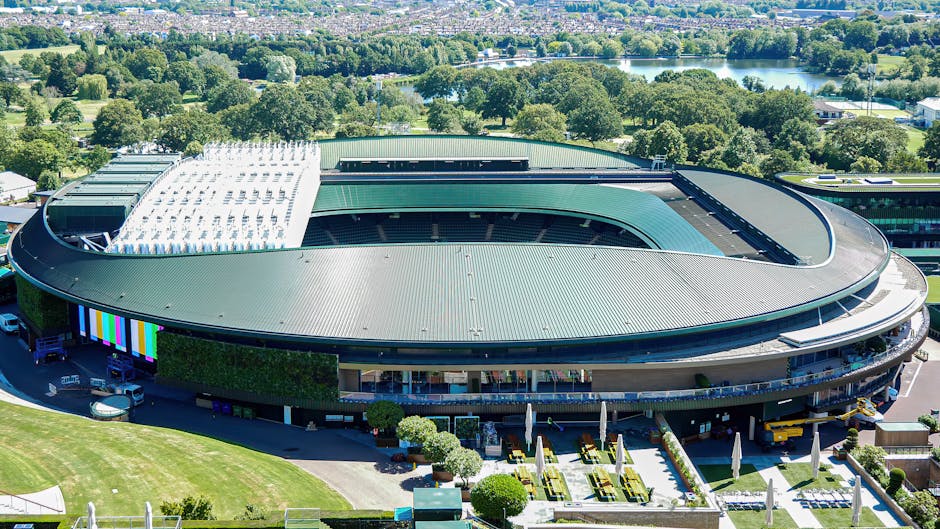 An aerial view of a large, modern racecourse stadium featuring a curved, elongated roof with a combination of smooth green and white roofing materials. The roof covers a central oval-shaped track area, visible through an open section in the roof's center, which appears to be an outdoor racing or sporting space. Surrounding the stadium, there are landscaped gardens, with neatly trimmed bushes and small trees, and outdoor seating areas with umbrellas. The stadium's exterior walls are primarily glass with some greenery vertical walls integrated into its facade. Multiple vehicles, including vans and service trucks, are parked along the curved driveway at the front of the building, which is connected to a spacious parking lot. The background includes lush green trees, open fields, and a lake, indicating a location within a park or countryside setting. The overall scene is bathed in natural daylight, emphasizing the stadium's clean lines, textured roofing surfaces, and well-maintained grounds, reflecting a setting where private event planning and venue hire may include on-site waste management options provided by local or private waste removal services such as Rubbish Removal Wimbledon.