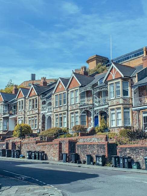 A suburban scene featuring a row of traditional terraced houses with bay windows, situated behind a black metal fence. In the foreground, there are two black waste collection bins positioned on a small patch of grass beside a paved footpath. A large, leafy green tree extends its branches across the upper left corner of the image, providing partial shade. The sky above is partly cloudy with patches of blue visible through the clouds. The environment appears well-maintained, and the setting suggests a residential area where private rubbish collection services, such as those provided by Rubbish Removal Wimbledon, might be employed for waste management on site.