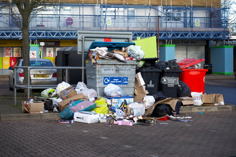 A collection of overflowing rubbish bins and scattered waste consists of various materials, including cardboard boxes, plastic bags, paper, and packaging, piled around and on top of a large grey mixed waste container situated on a paved urban street. The grey bin, labelled for mixed paper and cards, is open with crumpled paper, cardboard, and small packaging spilling out onto the ground. Adjacent to it are black and red refuse bins, also filled with waste, some tied in black bags, contributing to the cluttered scene. Surrounding the bins, loose items such as flattened cardboard, plastic food wrappers, and a few miscellaneous discarded objects are strewn across the pavement, indicating a state of neglected waste disposal. Behind the waste collection area, a multi-storey building with scaffolding, metal fencing, and a commercial frontage with signs can be seen, suggesting a busy commercial or residential district. The scene captures a typical instance of private waste accumulation that Rubbish Removal Wimbledon might address through independent rubbish collection services, emphasizing the importance of timely waste management.