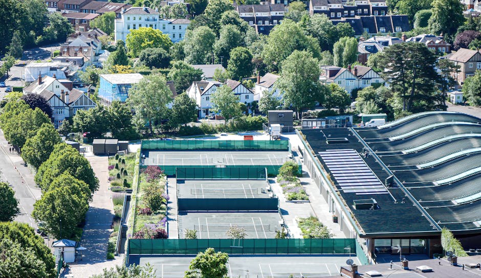 An aerial view of a mixed-use urban area showing a large commercial building with a distinctive curved black metal roof and multiple skylights, adjacent to a parking lot with several tennis courts enclosed by green fencing, and a surrounding residential neighbourhood filled with trees and detached houses with varied roof lines and colours. The scene is set in bright daylight with shadows cast by trees and structures, illustrating an area where private waste collection and alternative rubbish removal services, such as those offered by Rubbish Removal Wimbledon, might serve commercial and domestic clients in need of efficient rubbish clearance from both indoor and outdoor environments.