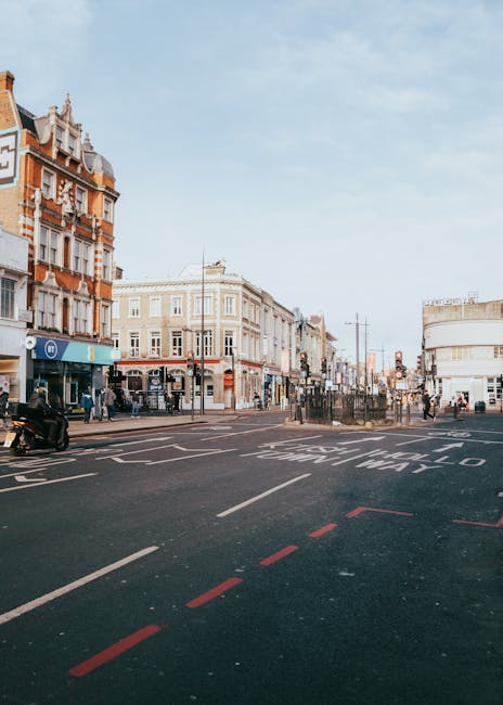 An urban street scene in Wimbledon featuring a wide paved road with clearly marked traffic lanes, including a dashed red line indicating parking restrictions or designated loading zones. On the left side, there are historic brick and stone buildings with ornate architectural details, housing various shops and businesses, with pedestrians walking along the sidewalks. To the right, a modern white commercial building with rounded edges and large windows contrasts with the older structures. In the background, a small group of people waits at a pedestrian crossing, while other individuals walk or stand further along the street. Traffic lights and street signs are visible, with a motorcycle parked on the sidewalk. The sky above is clear with a few light clouds, and the overall atmosphere suggests an active shopping or commercial area, where independent waste disposal and rubbish removal services, such as those provided by Rubbish Removal Wimbledon, are often utilized for maintaining cleanliness and managing waste in busy urban settings.