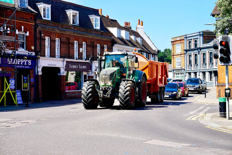 A large green and black tractor with a prominent front grille and large rugged tires is positioned in the middle of an urban street, attached to an orange waste collection container at the rear. The vehicle is situated near a row of mixed-use buildings, including a sports bar, a salon, and other shops, with a variety of shop signage visible. The street scene includes parked cars lining the right side, a traffic light showing red to the right, and a pedestrian crossing markings on the asphalt. Bright daylight illuminates the scene, casting shadows on the pavement and highlighting the textures of the brick and painted facades of the surrounding buildings. The environment suggests a busy town centre where private waste collection services, such as those provided by Rubbish Removal Wimbledon, might operate for building or commercial rubbish clearance. Details like the waste container and the vehicle’s purpose align with private onsite clearance activities for rubbish removal in urban settings.